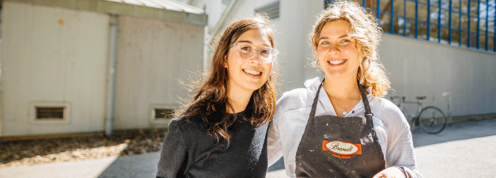 Photo of two female students outside a building wearing painting clothes and carrying a canvas